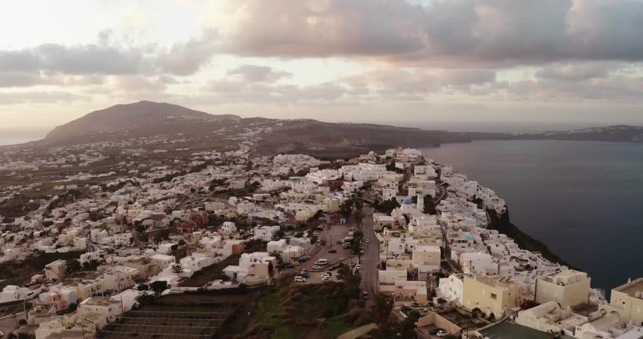 Wide drone view of the iconic white Cycladic village on the volcanic hillside of Santorini, Greece. The town cascades down toward the caldera and the sea, under a dramatic, cloudy sunset sky