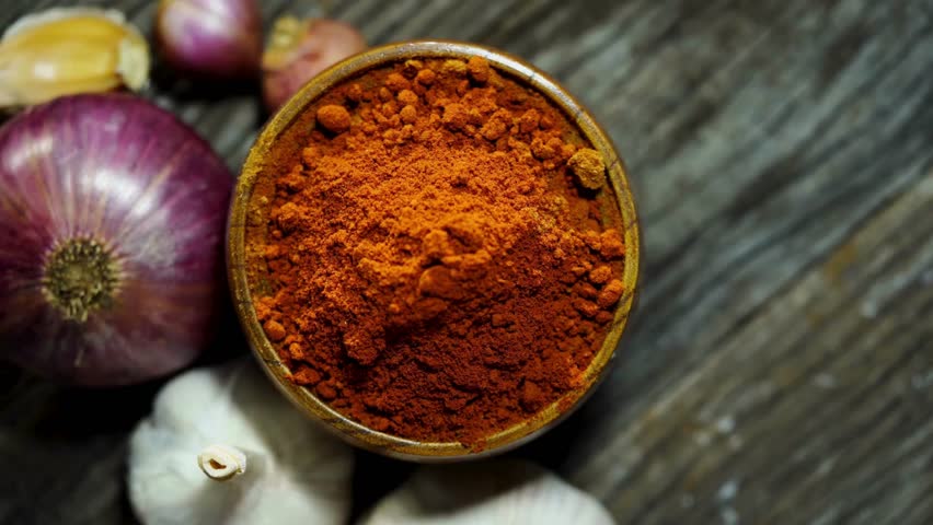 A bowl of red chili powder surrounded by onions and garlic on a rustic wooden surface.
