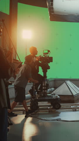 Vertical Screen: Professional Film Crew Records a Dynamic Action Sequence on Movie Set with Large Green Screen While Camera Operator and Cinematography Director Guide a Tracking Camera