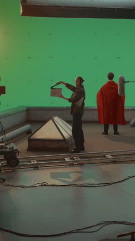 Vertical Screen: Behind-the-scenes Filming Superhero Actor in Red Cape on Green Screen While a Diverse Film Crew With Dolly Camera. Visual Effects Blockbust Movie Filmed on Controlled Studio Set