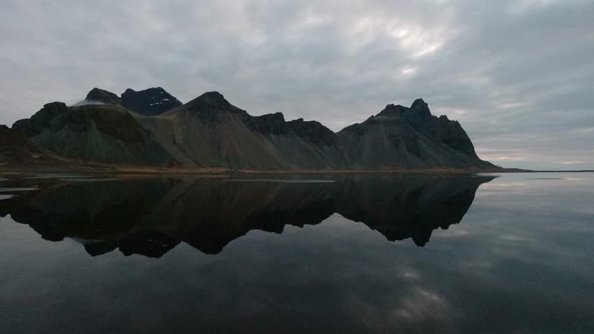 Static wide shot capturing the dramatic, jagged peaks of the Vestrahorn mountain reflected perfectly in the still, mirror-like dark water under a rapidly passing cloudscape.