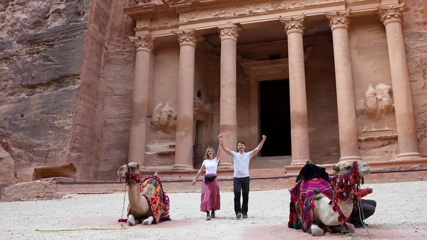 Traveler couple holding hands walk between two camels in front of famous Petra