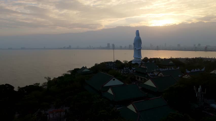 Breathtaking aerial drone view of the Son Tra Peninsula coastline at sunset, featuring the white Lady Buddha statue overlooking the ocean and Da Nang city