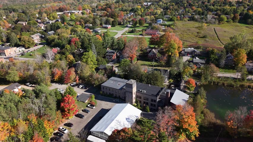 Drone shot of the Alton Mill Arts Centre in Caledon, Ontario, surrounded by fall foliage, heritage buildings, winding roads, and the pond reflecting autumn colours across the historic village.