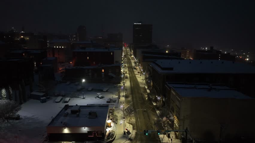 Winter dawn aerial over Lynchburg, Virginia, showing quiet downtown street lined with historic brick buildings, soft snow cover, warm streetlights and calm early morning atmosphere in United States.