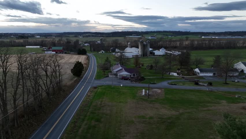 Aerial flyover of rural American open farmland, winding country roads, classic barns, grain silos, and scattered single-family farmhouses across wide, peaceful landscape at dusk. New England. USA.