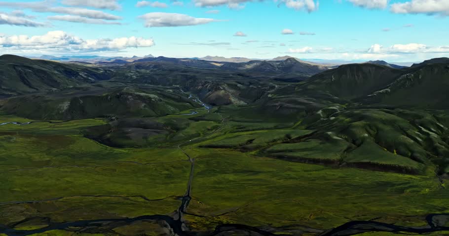 Expansive aerial view revealing a wide Icelandic valley framed by dark volcanic hills, distant peaks, and winding river channels under bright summer skies.