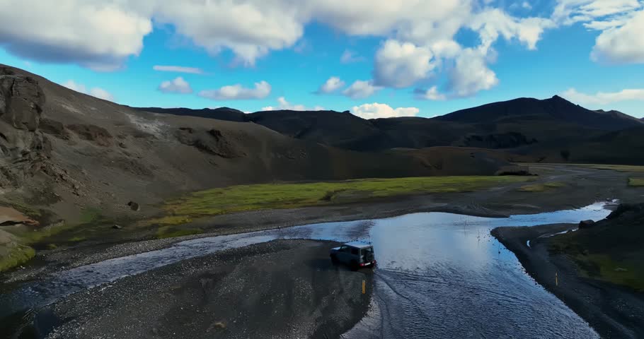A 4×4 navigates a shallow river crossing in the Icelandic highlands, driving toward a wide valley of dark volcanic slopes under bright summer clouds.