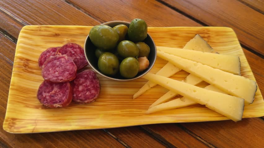 Wooden plate with a set of snacks for beer or wine Spanish sausage fuet, pieces of cheese, and green olives. High quality 4k footage