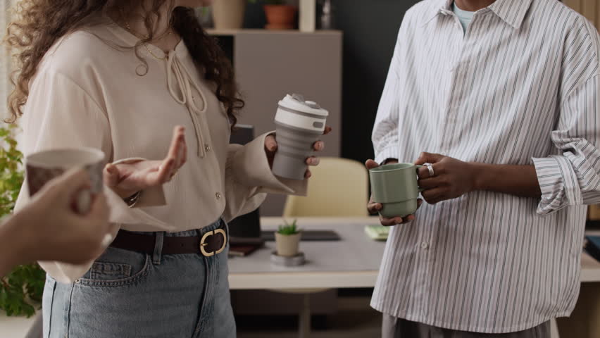 Three unrecognizable biracial coworkers standing together, holding cups full of coffee and enjoying conversation