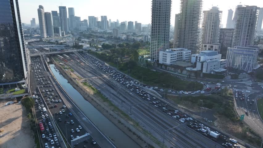 Aerial drone shot of the Ayalon Highway in Tel Aviv, featuring the central railway tracks, the Ayalon river canal, and busy traffic flowing between modern high-rise buildings.