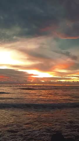 A sunset on the beach with a golden-orange sky and dark clouds framing the horizon. Gentle waves lapping against the shoreline, reflecting the warm, serene light of dusk. 