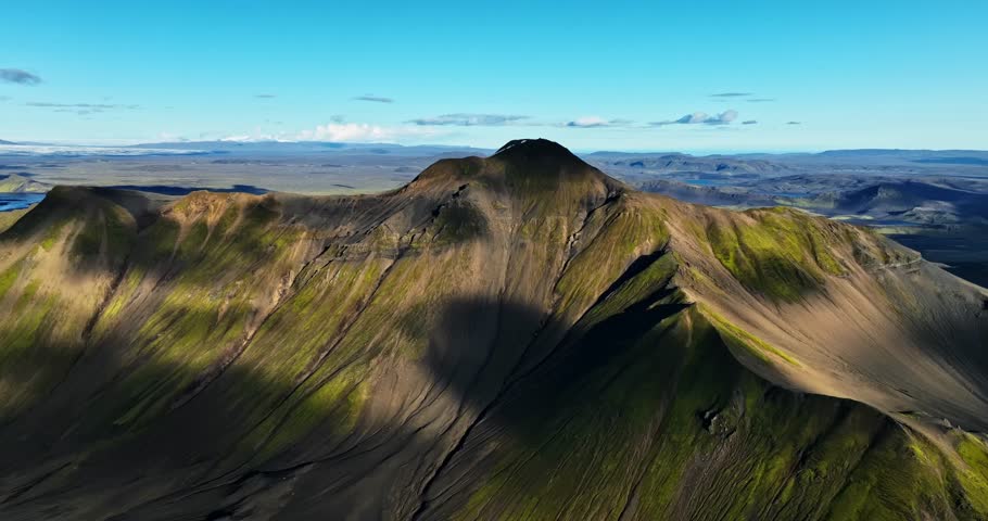 High drone angle revealing a steep volcanic ridge lit by afternoon sun, exposing rugged textures, mossy slopes, and a vast Icelandic plateau stretching endlessly into the horizon.