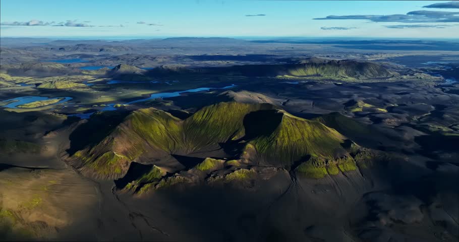 Drone view over Iceland’s dark volcanic plains and moss-covered craters, revealing layered ridges, distant lakes and long evening shadows across the highlands.