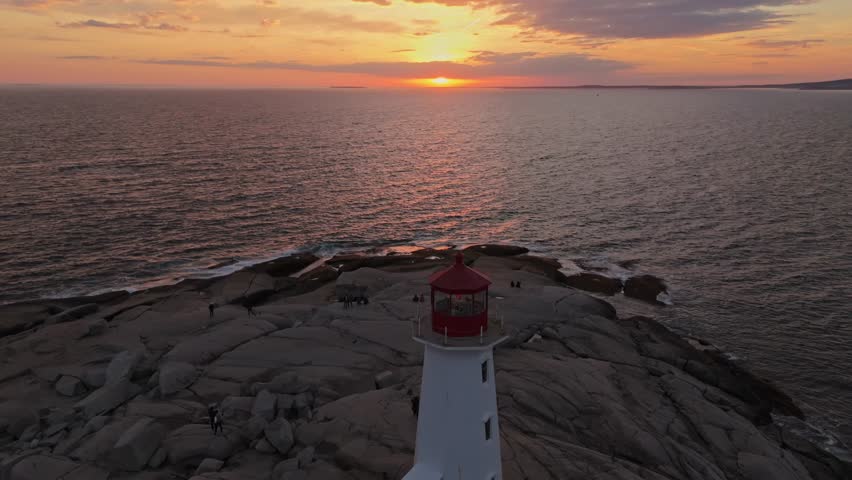 Cinematic Aerial View Of Peggys Cove Near Halifax, Nova Scotia, Featuring The Iconic Lighthouse Standing On Massive Granite Boulders During Sunset, With Soft Reflections Across The Shoreline
