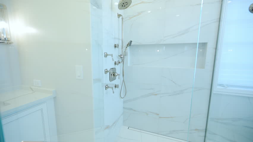 Clear glass shower door in a bright, modern bathroom. The background reveals white cabinetry, polished marble tile, and high-end shower fixtures