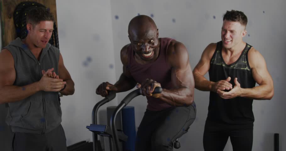 Main rider pedaling air bike, fitness partners clapping and urging, dotted spiral appearing behind. Gym, workout, training, cardio, motivation, encouragement, teamwork