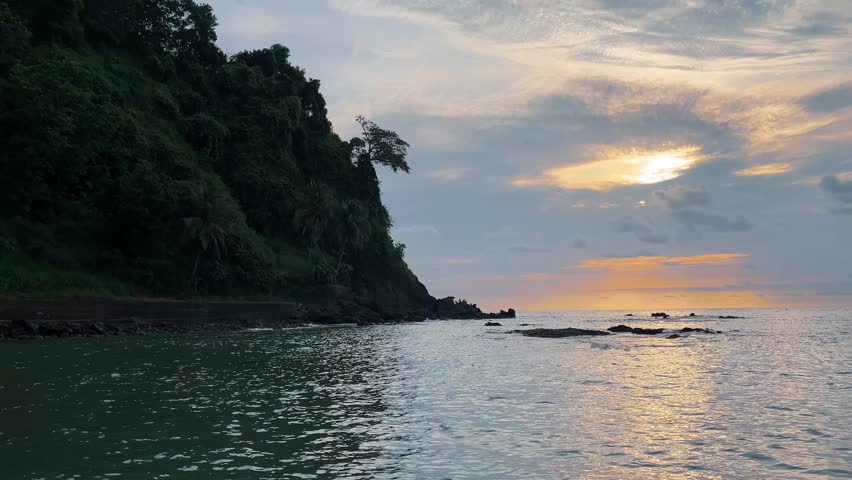 a peaceful coastal scene featuring a lush green cliff beside the ocean. gentle waves hit the rocky shoreline as the sun sets behind scattered clouds