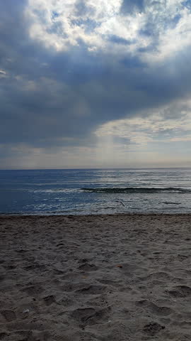 Large dark rainy clouds in sky over sea. Sandy beach on seashore and rays of light shining through clouds on summer day. Sea, sky, clouds, horizon line. Rain in sea. Seascape. Sea Landscape. Vertical