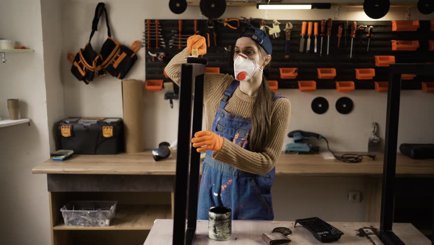 Woman in respirator mask painting metal table legs at workshop. Craftsman furniture