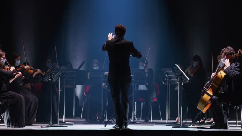 Cinematic shot of a conductor leading a symphony orchestra on a classic theatre stage, with musicians performing violins, cellos, and trumpets during a live concert.