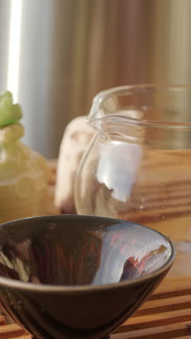 Close-up of boiling water being poured from clay teapot into glass pitcher and ceramic tea bowl set on wooden tea tray. Warming up tea brewing utensils. Traditional chinese gongfu tea ceremony at home