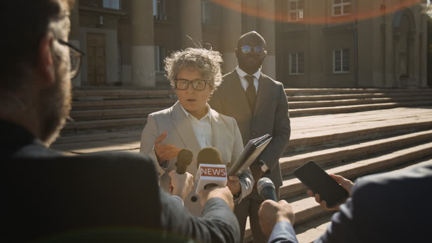 Medium shot of mature Caucasian female criminal defense attorney standing on steps of court hall, speaking to journalists and giving expert comments on case, African American security detail watching