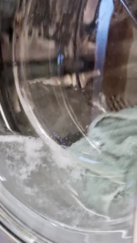 Vertical, close-up front-load view inside a modern washing machine drum. Laundry is agitating and tumbling in foamy water with bubbles, with green and brown fabrics rotating through a door.