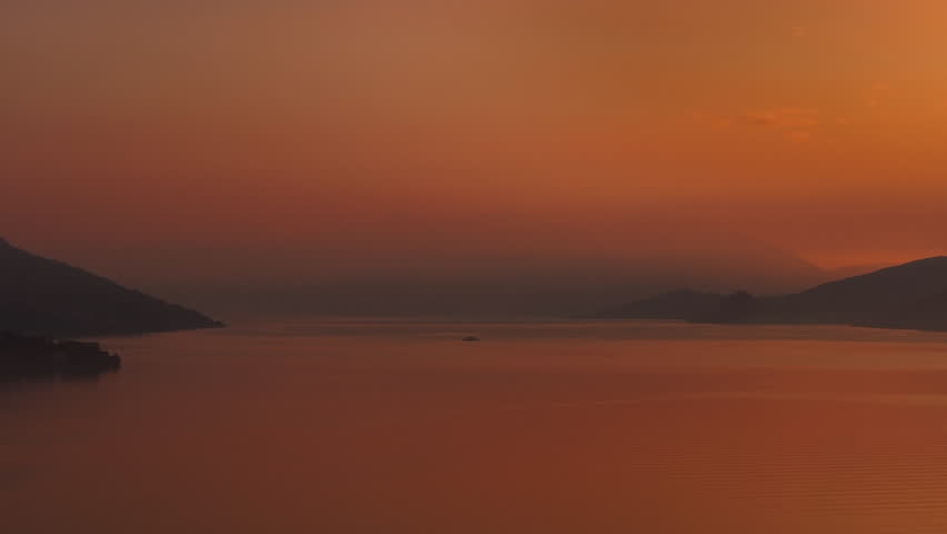 deep orange dusk over quiet coastal bay soft gradients, calm water with subtle ripples, distant islands and empty horizon, serene