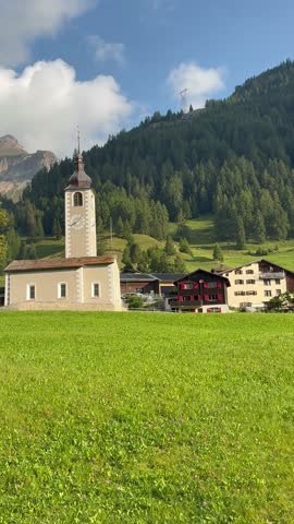 Sufers, Switzerland. Historic village church and chalets in a sunny, green Alpine meadow. The view includes a dramatic mountain panorama of rugged peaks under heavy storm clouds.