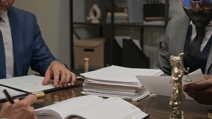 Medium shot of Caucasian and African American male lawyers discussing legal precedent, while reviewing case files during team meeting in office, working on defense arguments