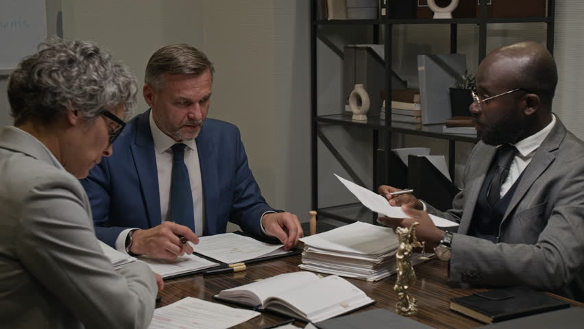Medium shot of three diverse attorneys reviewing archive of case documents while preparing for court case, African American lawyer showing legal precedent to senior partner and proposing strategy