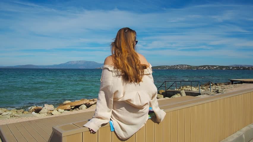 A girl sits with her back to a wooden equipped beach island for vacationers near a rocky beach, with the sea and mountains in the background. Aegean coast, Cesme, Türkiye