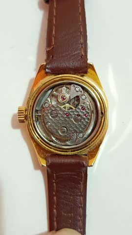 Close-up video of a mechanical wristwatch with moving balance wheel, filmed from the open back case, featuring a golden watch body and brown leather strap, showcasing precision craftsmanship.