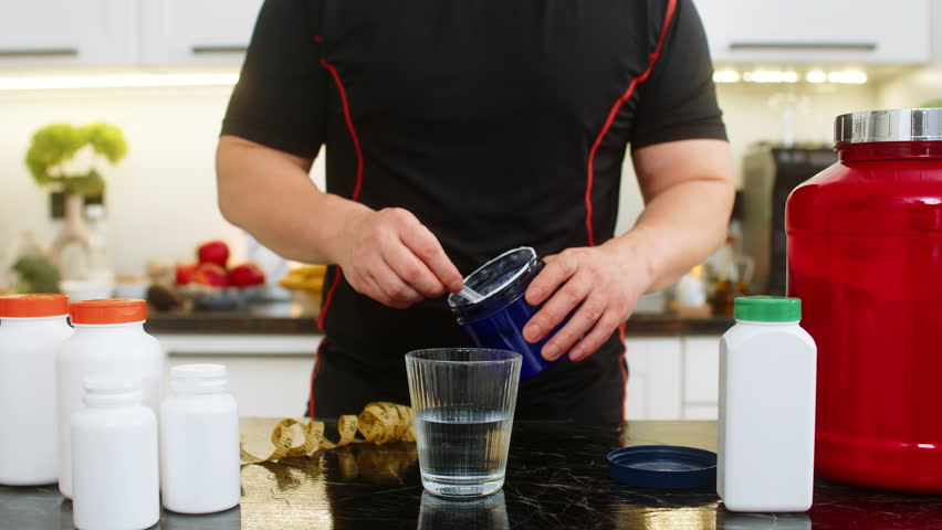 Sportsman pours scoop of supplement powder into glass of water in home kitchen. Concept of mixing electrolytes isotonic creatine glutamine bcaa slimming drink for fitness energy and muscle recovery.
