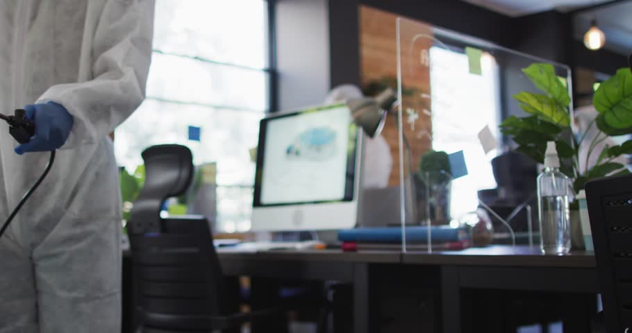 Worker entering from left disinfecting office, spraying plexi while DNA overlay obscuring monitors. Sprayer, sanitization, hazmat, gloves, ppe, acrylic, workspace