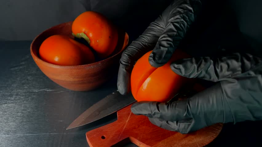 Hands in black gloves holding halved juicy persimmon. Fresh fruit on cutting board, wooden bowl with whole persimmons in background. Natural produce, healthy seasonal food, organic raw food.