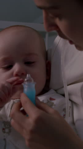 Mother gently cleans baby's nose with a nasal aspirator while seated in a cozy nursery, showcasing the tender interaction, camera pans to capture the moment