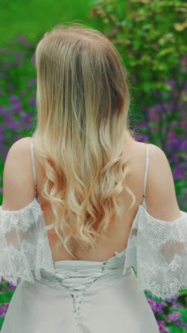 young woman enjoy summer green nature flowers spring garden rear view. lady straightens long healthy curly blond hair with hands. white lace dress old style open bare back. adult girl, vertical video