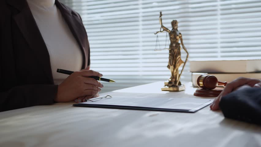 Professional female lawyer explaining legal document terms to a client during a consultation. A gavel and scales of justice statue sit on the desk