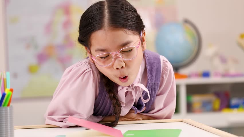 Asian schoolgirl in pink glasses clutches stomach in pain at bright classroom desk