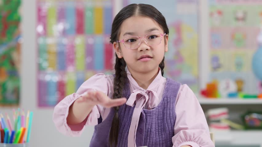 Young student with glasses eagerly raises hand in brightly lit classroom, holding notebook, mid-discussion