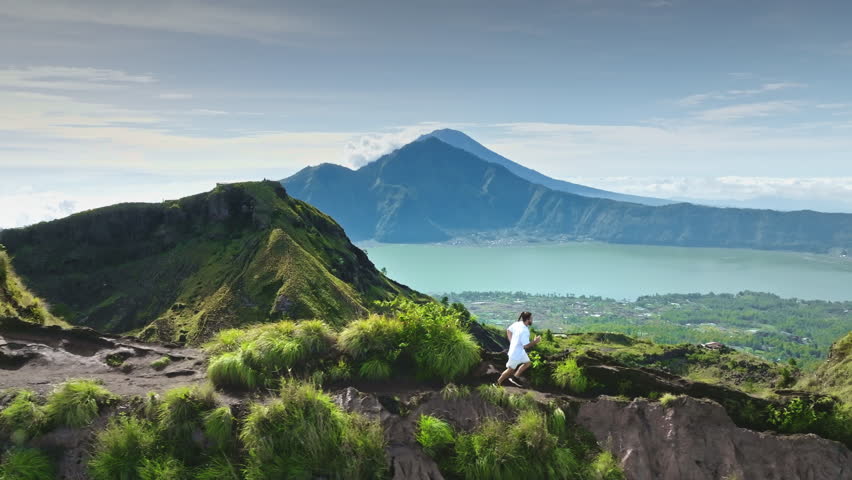 Man running on mountain trail, actively training and exploring rugged volcanic terrain with Lake Batur and Mount Agung in the background, sports, adventure, health, and travel. Aerial drone flight