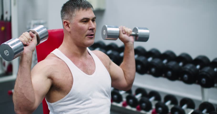 Seated bench press with dumbbells for the deltoid muscles. Deltoid workout with dumbbells. Slow motion 4k footage of a middle aged brutal Caucasian man during gym workout.