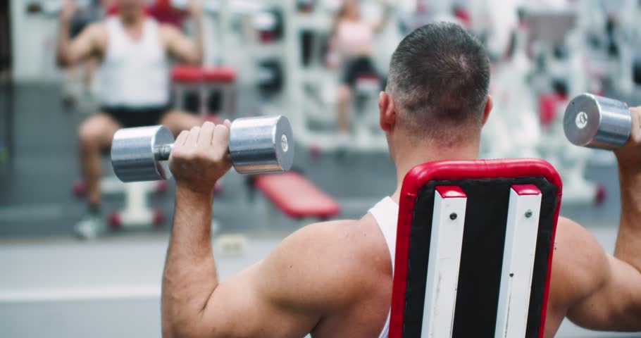 Seated bench press with dumbbells for the deltoid muscles. Deltoid workout with dumbbells. Back view of a middle aged brutal Caucasian man during gym workout in front of a mirror. 4k footage.