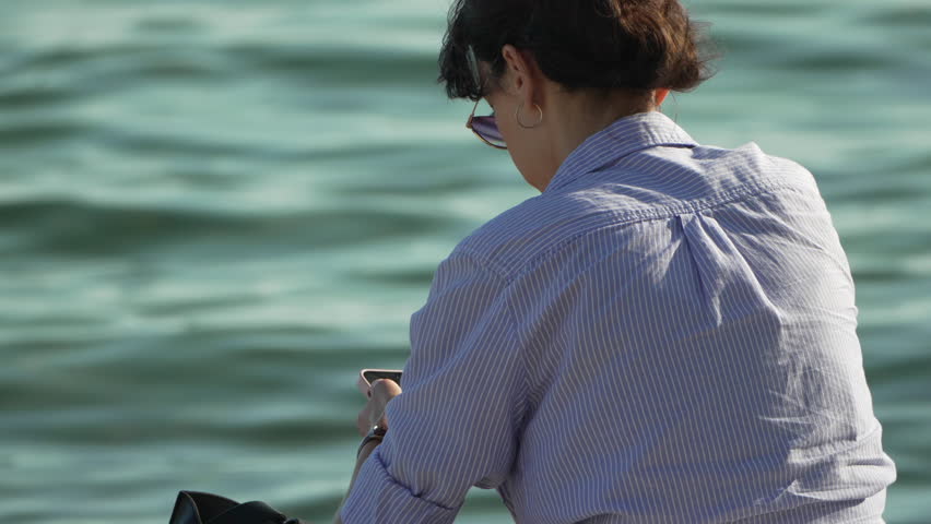 Woman, smartphone, water: Relaxed woman using phone by calm water on a sunny day.