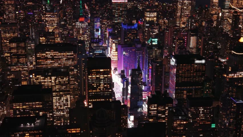 Aerial close up view of Times Square and surrounding Midtown Manhattan skyscrapers at night. Buildings are illuminated with bright electric and neon signs