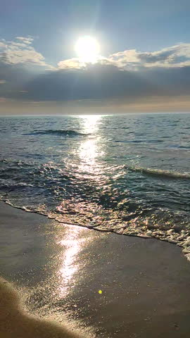 Seascape on sunny summer day. Sea waves rolling on sand of sandy beach on seashore. Waves of calm sea, sand, blue sky with clouds and horizon line. Sun shining from behind rainy clouds. Vertical