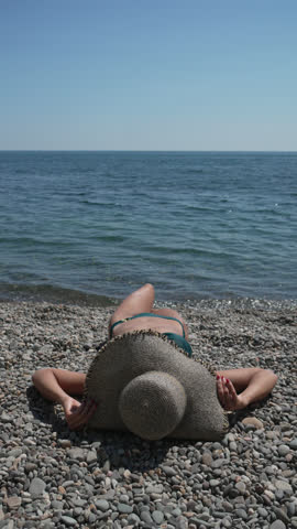 Woman beach relaxing. Young woman sunbathing on pebble beach by blue sea under clear sky, summer vacation. Vertical video.
