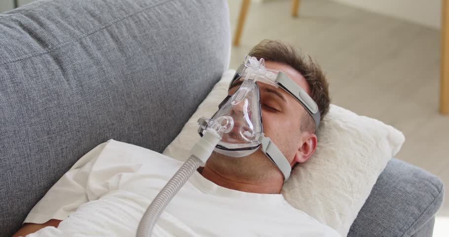 Man mask sleep apnea therapy oxygen CPAP. Adult patient rests on sofa using CPAP device for home healthcare and safer breathing at night. Clear medical concept of sleep apnea therapy.